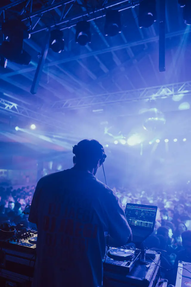 DJ in headphones seen from behind, facing a crowd under blue stage lights